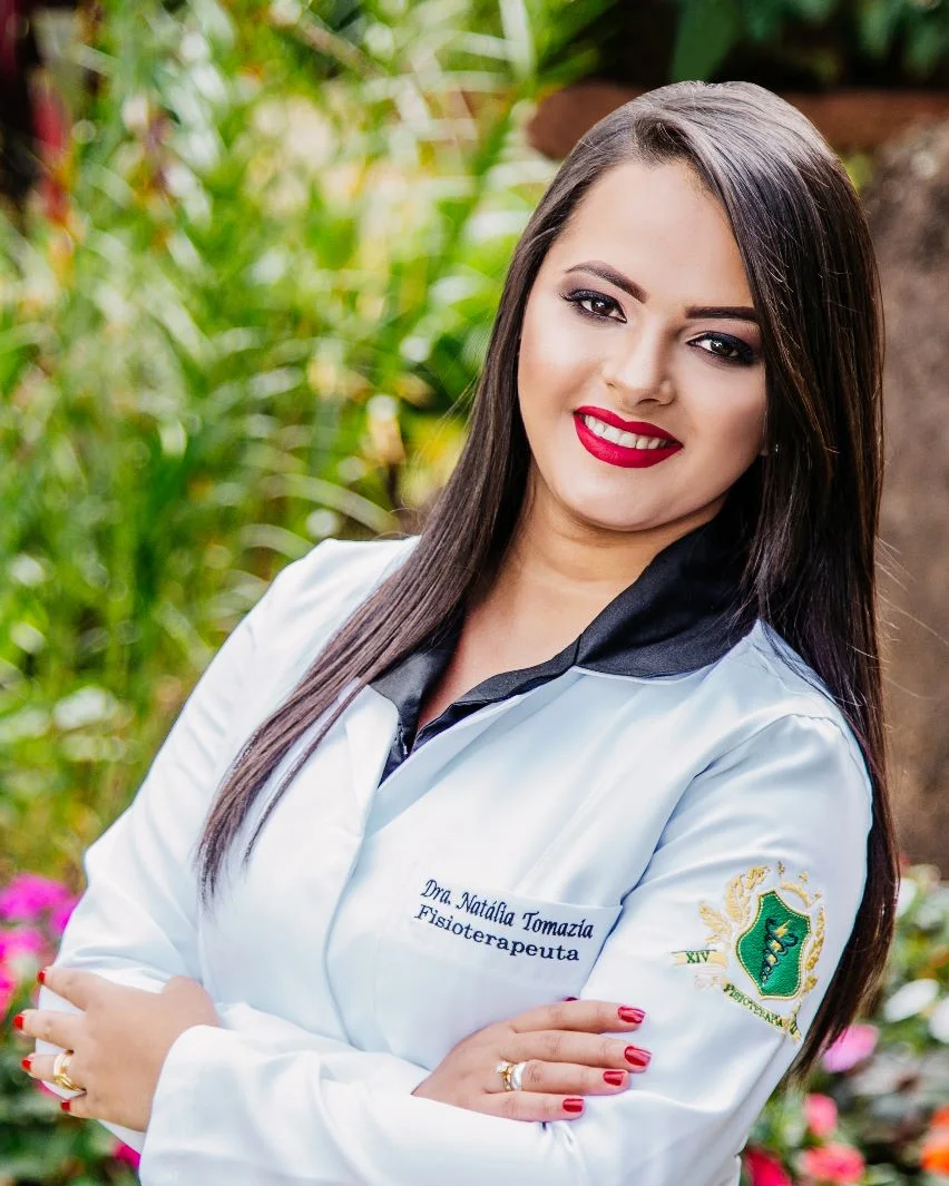 Natália Tomazia, fisioterapeuta, de jaleco branco e braços cruzados, sorrindo em um jardim